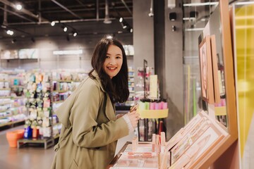Beautiful woman testing and buying cosmetics in a beauty store. Young girl trying lipstick on hand at cosmetic store. Retail and consumerism. Personal care and modern cosmetology. Side view