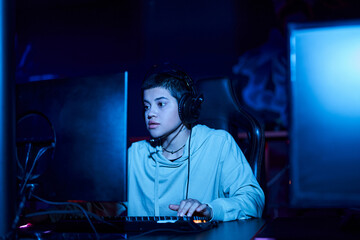 focused young player looking at computer monitor while playing game in a blue lit room, cybersport