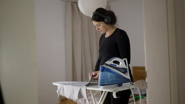Pregnant woman ironing clothes at home doing domestic chores while wearing headphones listening to content online, person multi-tasking
