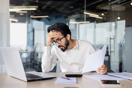Focused indian businessman in shirt working on laptop with paperwork at office desk