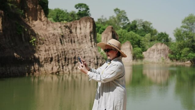 Portrait, beautiful elderly Asian woman, dress beautifully, wear clothes, sunglasses, large hat protect from sun from hot weather, standing taking photos near water's edge with small mountain.
