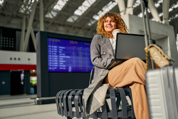 A woman freelancer, laptop in use, sits with a suitcase, waiting for her flight, enjoying a cup of takeaway coffee.