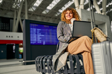 A woman freelancer, laptop in use, sits with a suitcase, waiting for her flight, enjoying a cup of...