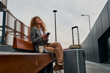 Sitting on a bench, a Caucasian woman uses her phone, holding a takeaway coffee with a suitcase...