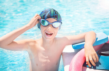 Active child (boy) in cap, sport goggles ready to learns professional swimming with pool board, swim noodles. Kid enjoying water in swimming pool. Healthy lifestyle.