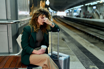 At the railway station, a tired woman sits, holding a suitcase, enduring a headache.