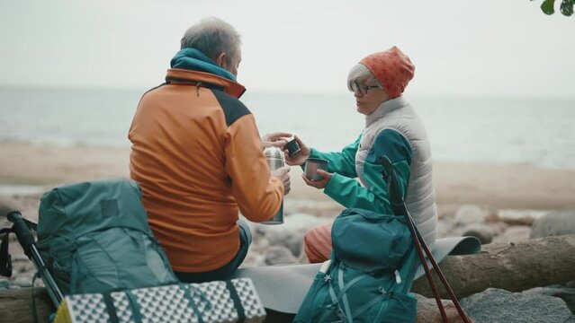 Senior man woman campers drinking hot drink from thermos talking enjoying nature on riverbank. Old couple family pensioners travelling together. Travel tourism camping journey hiking concept.