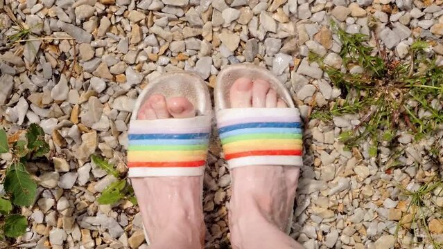 Water is pouring on female feet in rainbow sandals on pebbles, view from above. Summer sunny day.