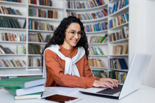 Smiling casual brunette typing on laptop keyboard while sitting in campus library with academical books aside. Promising graduate student finishing article and feeling satisfied with research result.