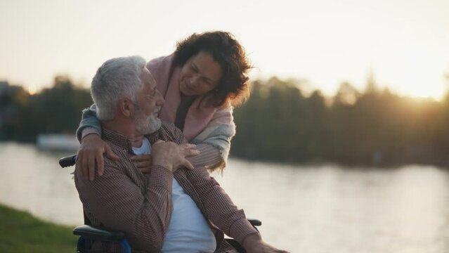 Wife walking with husband in wheelchair. Senior family couple spending time in city park near pond. Happy woman hugging man showing love care, they talking. Living with physical disability concept.