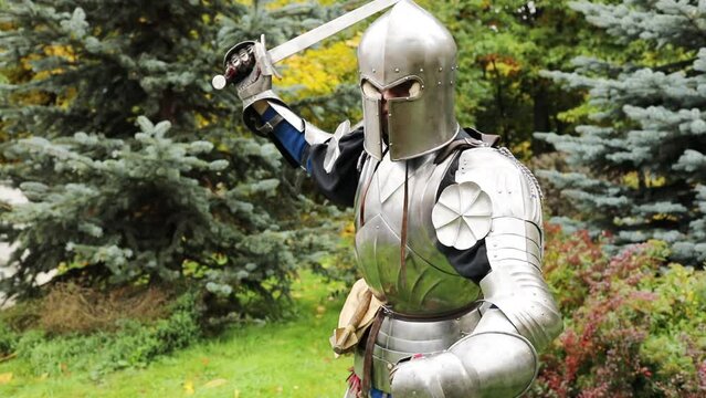 Close-up of man in medieval iron armor threatens with sword in hand standing on green grass, slow motion