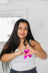 young latin brunette making a heart with her hands, with a pink ribbon symbol of the fight against breast cancer.