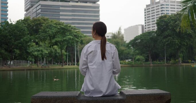 Woman sits on bench in Bangkok public park skyscrapers in background near water lake back view. Bangkok park blend of Thailand's urban and natural beauty Bangkok park tranquil and green.