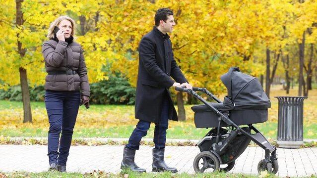 Woman talks by phone and man stands with baby carriage in autumn park, slow motion