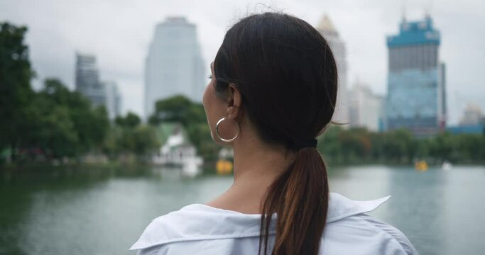 Back view of woman standing on park grass Bangkok's skyscrapers rising lake nearby. Tranquility in urban outdoors Tranquility blending cityscape with nature Tranquility peaceful amidst city.