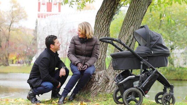 Happy man and woman talk near tree and baby carriage near river in fall park