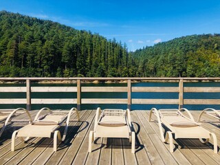 Empty sunbeds on a wooden pontoon near a lake surrounded by green forest - obrazy, fototapety, plakaty