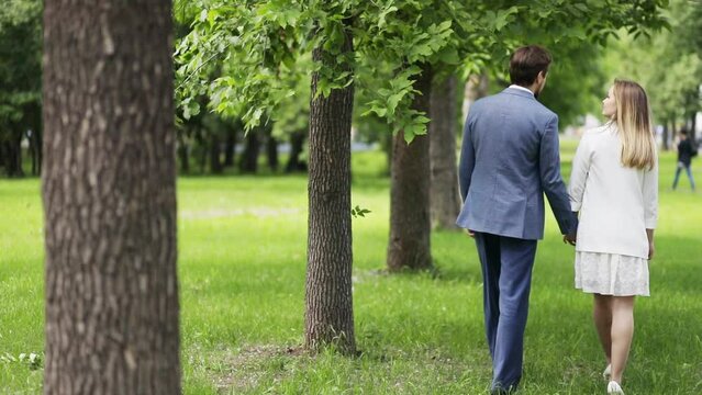 Happy pair walks near green trees in city garden at spring day, slow motion, back view