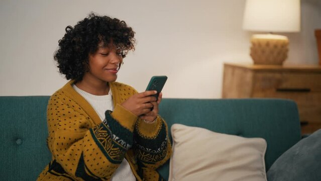 Modern woman with phone talking with friends. African American girl hold in hand use mobile phone. Teenager relax spend free spare time in living room with phone. Communication on social networks.