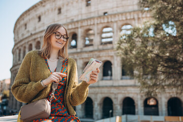 A happy blond woman tourist is standing near the Coliseum, old ruins at the center of Rome, Italy. Concept of traveling famous landmarks. Girl with map is walking on a sunny day