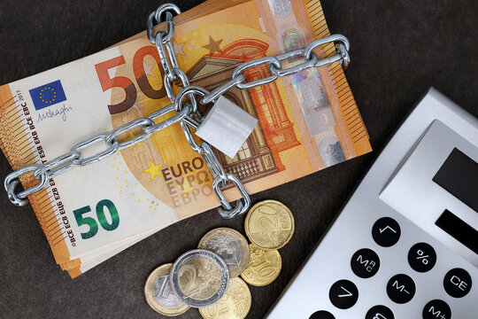 A stack of cash with 50 euro banknotes is locked with a chain and padlock next to a calculator and coins, concept for the safe investment of fixed-term deposits or financial security in the future