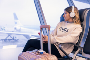 Relaxed attractive caucasian young woman using a smartphone at the airport with suitcase next to her and headphones looking at the window