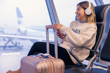 Relaxed caucasian young woman laughing while using a smartphone at the airport with suitcase next to her and headphones