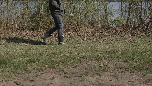 Strange and suspicious unrecognizable man in rubber boots walks through the wilderness. Slender male person keeps his hands in pockets and walks forward, side view.
