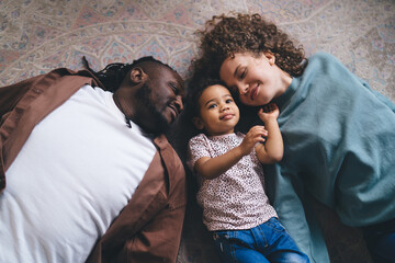 Happy multiracial parent with little daughter lying on floor together in living room