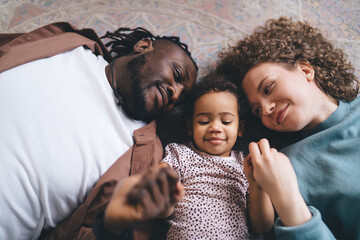 Top view of happy multiracial parent with little daughter lying on floor while playing together