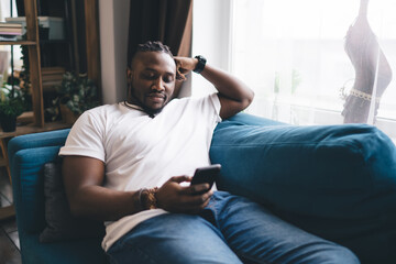 Positive black man browsing smartphone while sitting on sofa