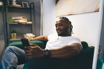 Positive black man browsing smartphone while sitting on sofa