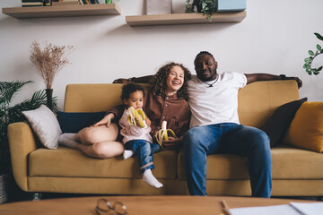 Cheerful diverse family eating banana while having fun together on sofa