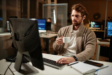 Portrait of bearded man using computer in office and drinking coffee while working late at night, copy space