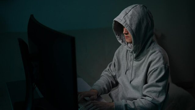 Tracking shot of hacker man in hoodie typing on keyboard breaking password using desktop pc sitting at desk in dark room. Dangerous hooded hacker breaks into data servers infects system with virus.