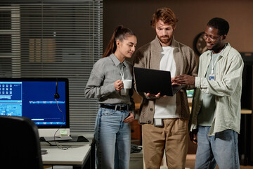 Waist up portrait of three young people using laptop standing in IT office at night, copy space