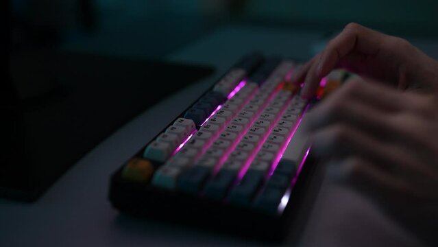 Detail cropped shot of unrecognizable man freelancer male typing on wireless RGB mechanical keyboard with colorful keycaps with ASMR sounds, sitting at desk in dark room. Shooting in slow motion.