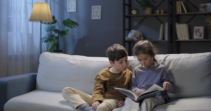 Children reading book, looking at pictures, sitting on the sofa in the evening. Leisure time. Childhood, education concept