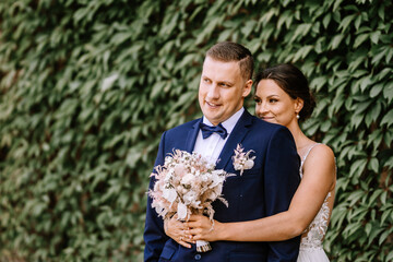Valmiera, Latvia - July 7, 2023 - Bride and groom are smiling, with the bride behind the groom and green leaves in the background.