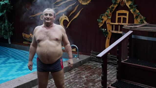 Vapour goes from old man which stands near pool after russian bath