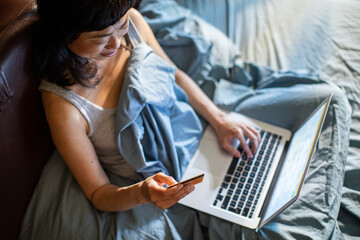 Happy woman using credit card on laptop in bed at home