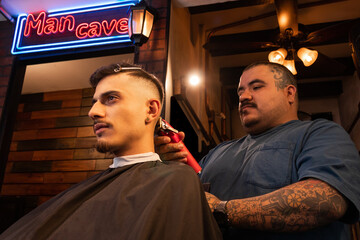 Tattooed Barber cutting hair of client with clipper in barbershop