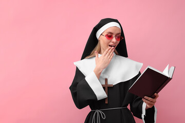 Surprised woman in nun habit and sunglasses reading Bible against pink background. Space for text