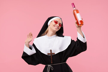 Happy woman in nun habit and sunglasses holding bottle of wine on pink background