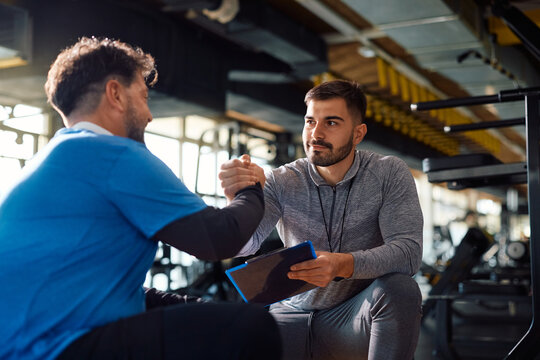Young coach gives manly handshake to mature man after successful sports training in gym.