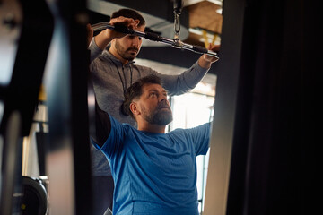 Personal trainer assists mature man in using exercise machine during gym workout.