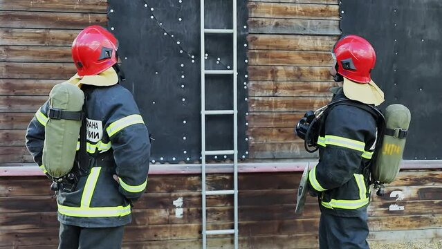 Two firemen in protective suits with russian inscription Fire Guard of Russia Emercom, stand near ladder on training site. Slow motion