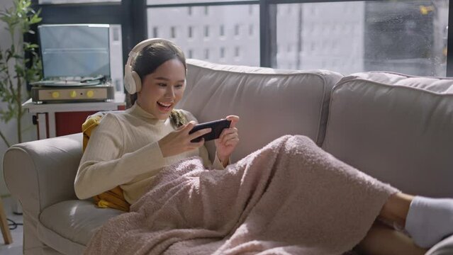 Beautiful woman using smartphone to play online video game on sofa in living room.happy young asian woman wearing headphones and playing game online with mobile phone on couch.