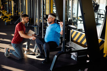 Fototapeta na wymiar Mature man working out on exercise machine during exercise class with personal trainer in gym.