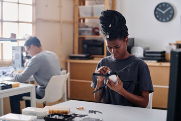 Young black technician repairing drone at her table in workshop, male coworker sitting on background - obrazy, fototapety, plakaty
